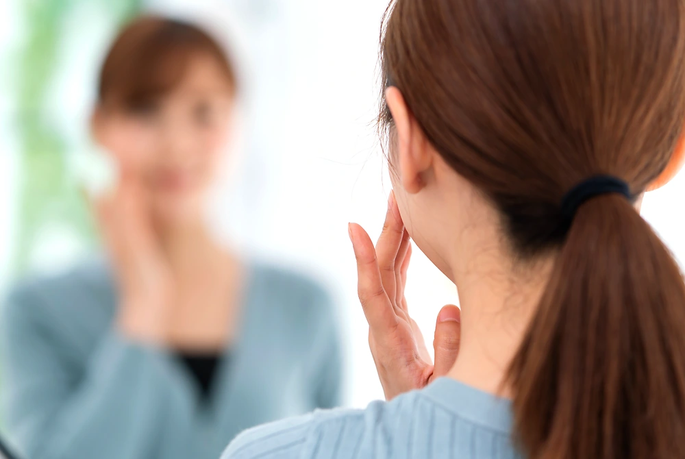 鏡の前で顔に手を添え、自分の外見を確認している女性。