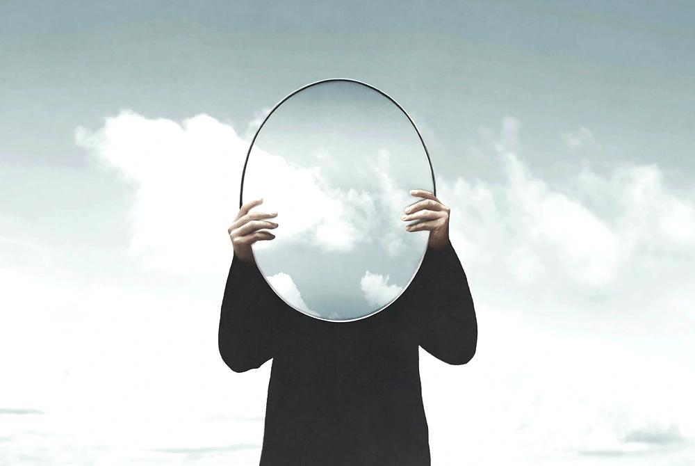 人物が顔の前に丸い鏡を持ち、鏡の中に雲が映っている抽象的な画像。