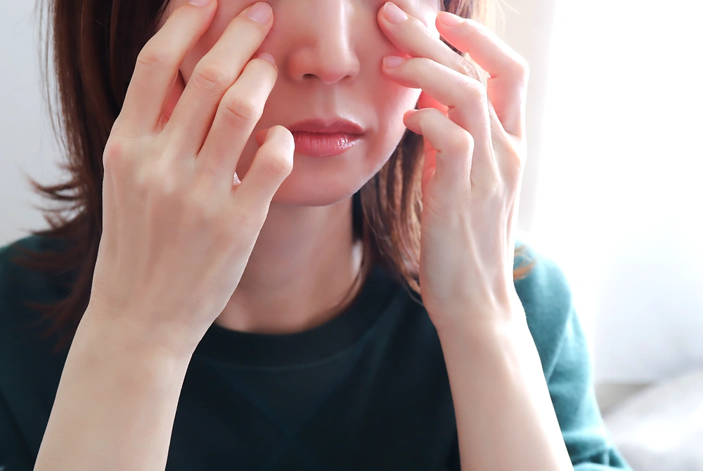 鏡の前で目の下に手を添え、下まぶたのふくらみやたるみを確認する女性。