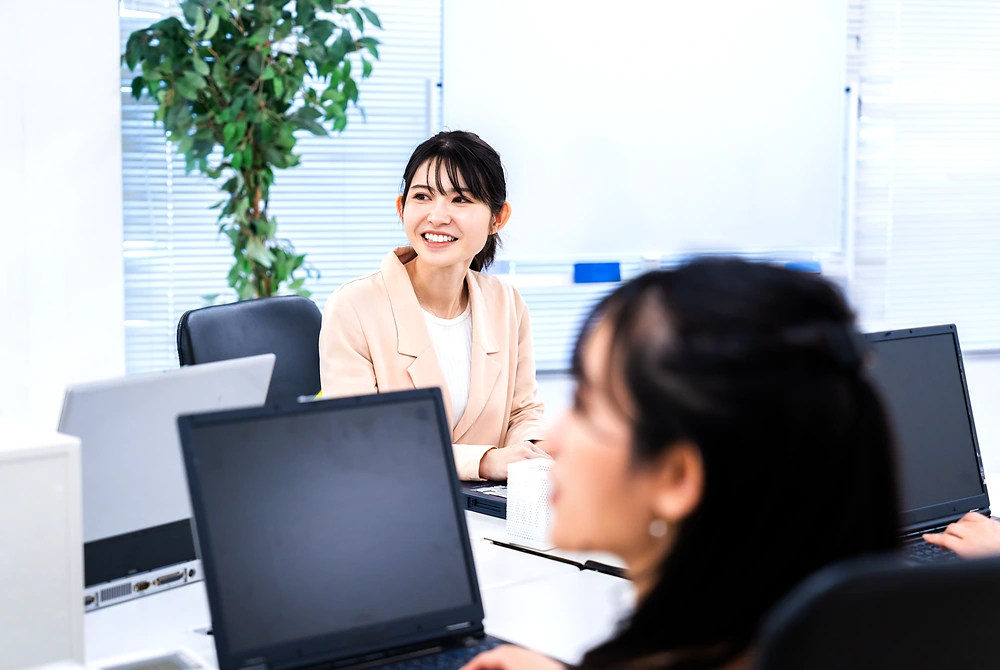 オフィスで複数の女性がパソコンを前に会話している様子。