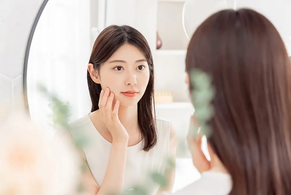 鏡の前で頬に触れながら肌の状態を確認する女性。