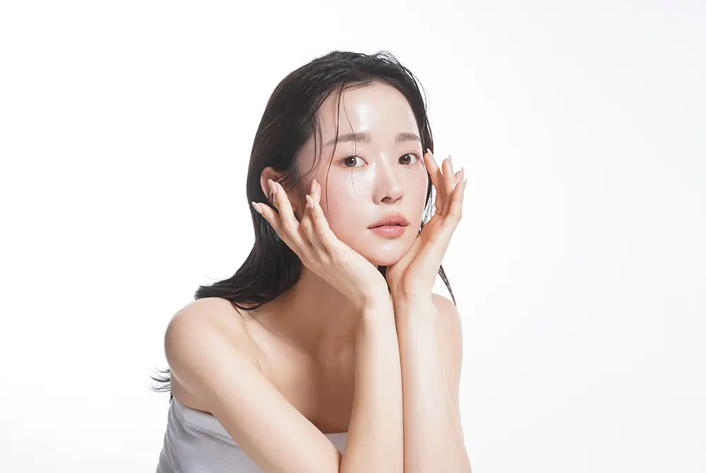 白い背景の前で頬に両手を添え、肌を見せる女性の美容イメージ。
