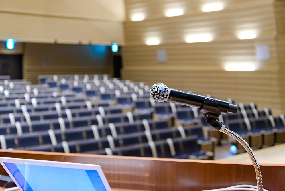 講演会場の演台に設置されたマイクと、奥に並ぶ空席の座席。