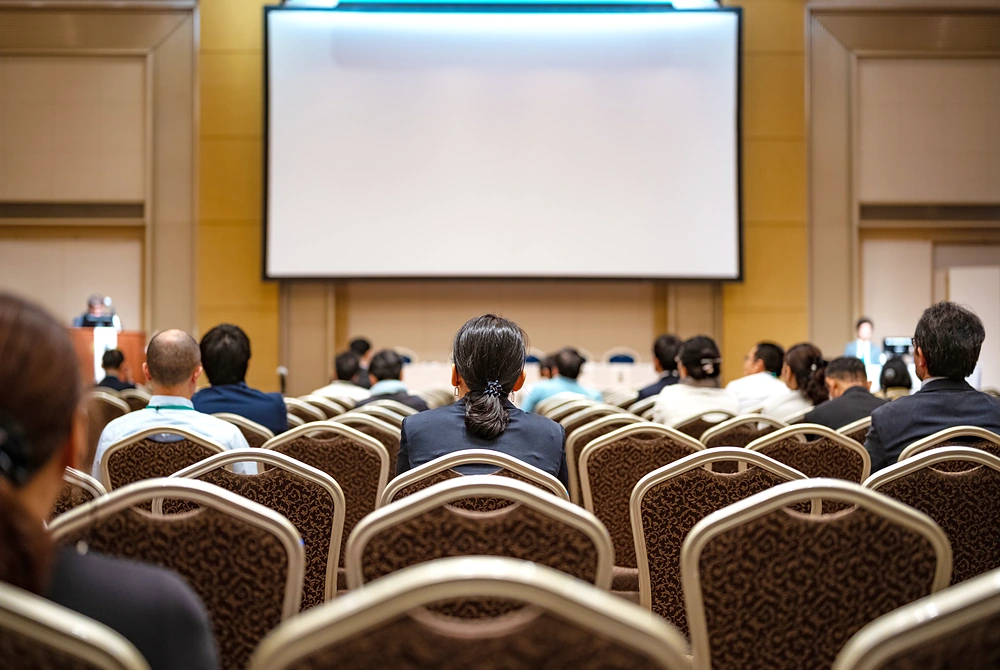学会や講演会の会場で、参加者がスクリーンに向かって座っている様子。