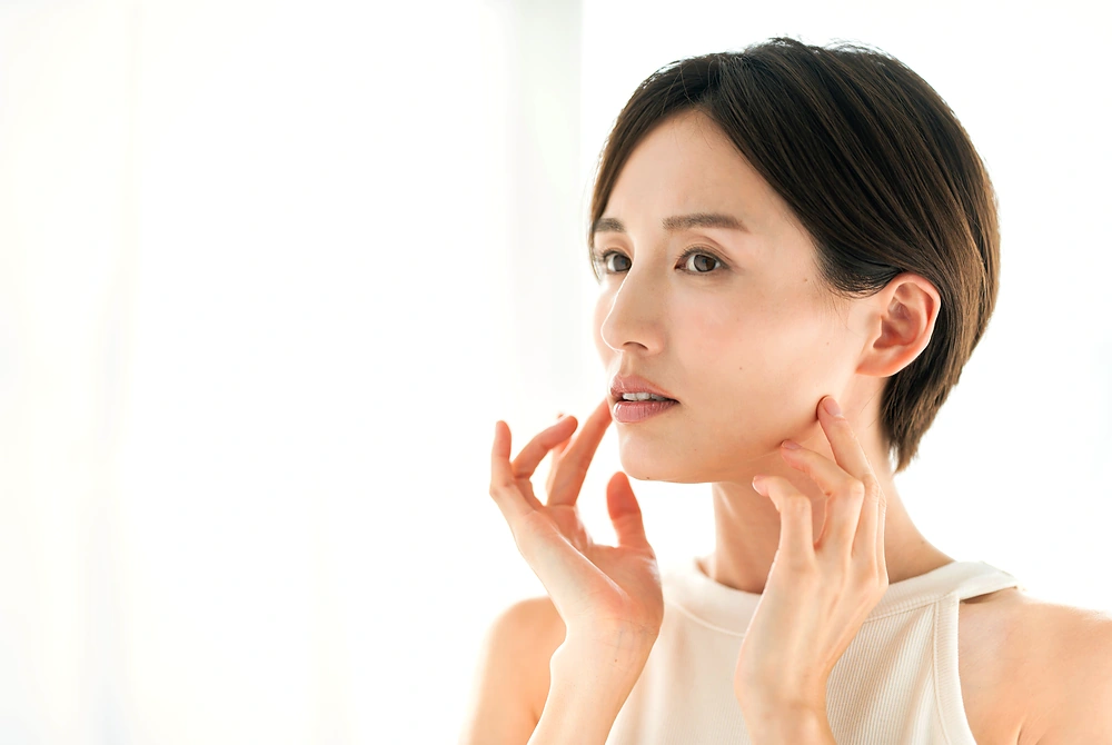 明るい室内で、頬やフェイスラインの肌に手を添えて状態を確認する女性。