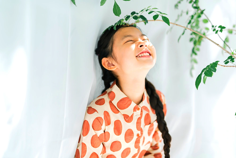明るい室内で、植物の葉の下で笑顔を見せる女の子。