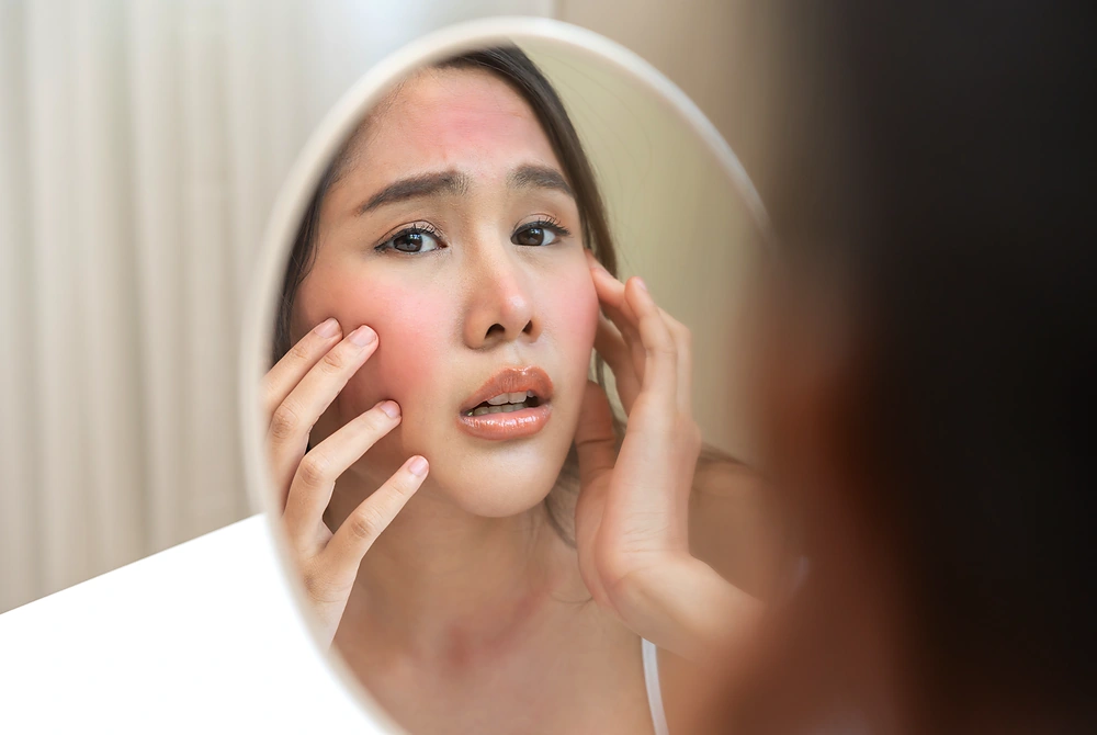 鏡を見ながら、赤みのある頬や額に触れて肌の状態を確認する女性。