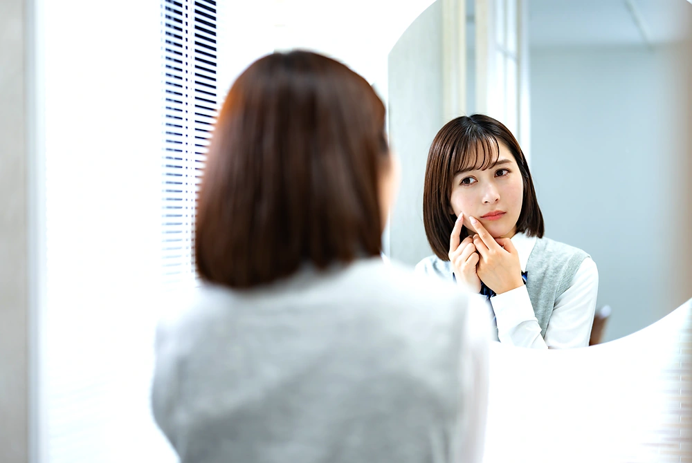 鏡を見ながら頬のニキビ痕や肌の凹凸を確認する女性。