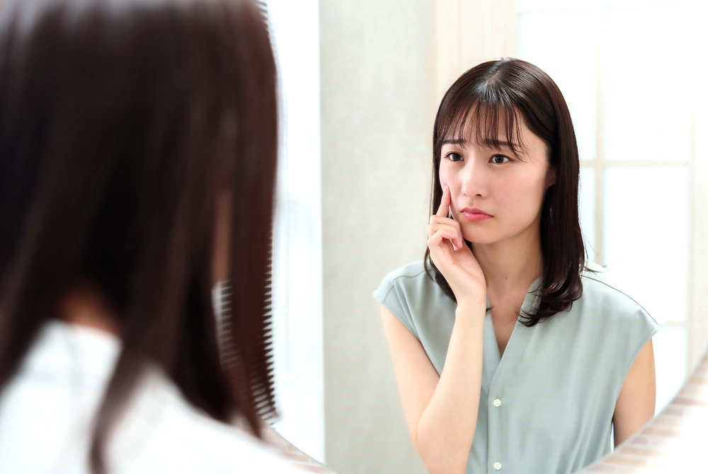鏡を見ながら頬のニキビ跡を確認する女性。