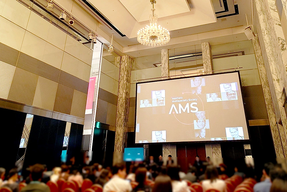AMWC Japan 2026の講演会場で、大型スクリーンに学会ロゴが映し出され、参加者が着席している様子。