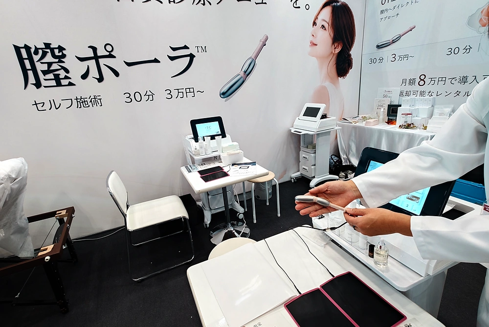 フェムケア機器を紹介する展示ブース。膣内ケアを目的とした機器や施術用ベッドが設置され、担当者が機器を手に持って説明している。